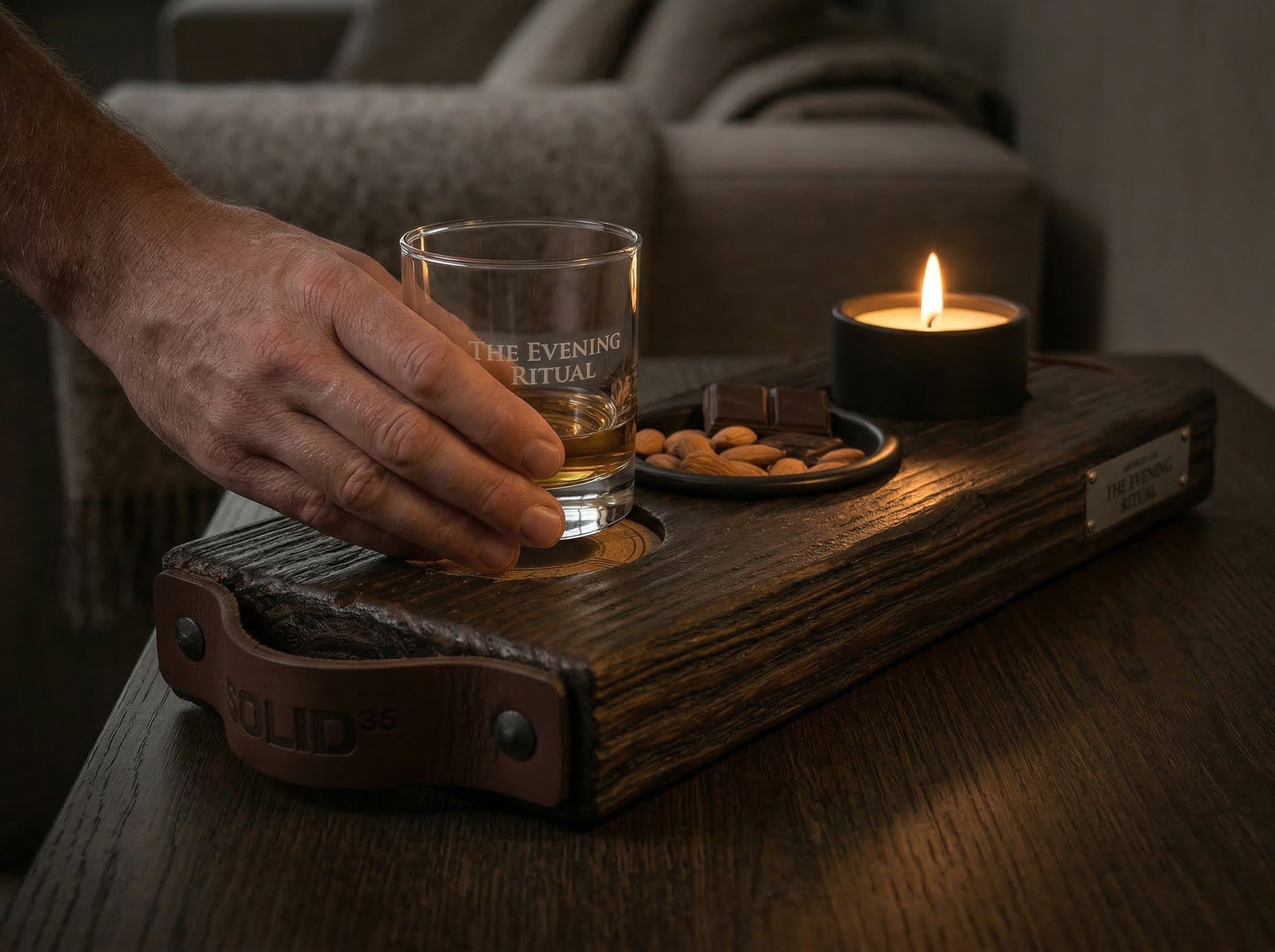 Hand pakt een gegraveerd whiskyglas van The Evening Ritual serveerplank met snacks en brandende kaars.