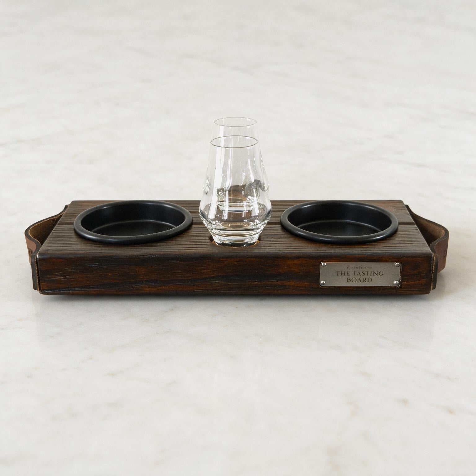 Vooraanzicht van The Tasting Board met twee proefglazen, zwarte schaaltjes, leren handgrepen en metalen naamplaatje
